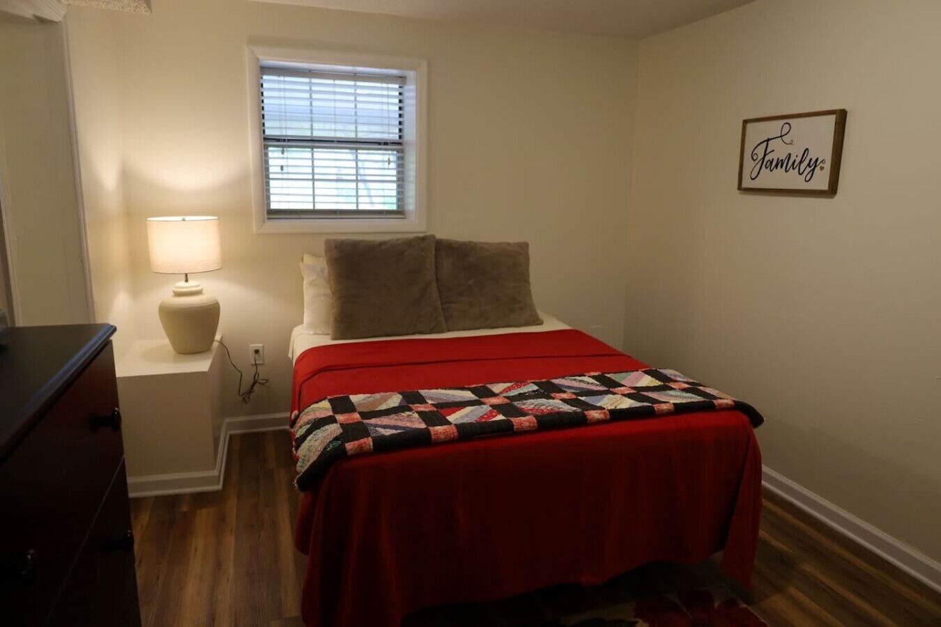 A bedroom with a bed, a nightstand, a lamp, a window with blinds, and a 'Family' sign on the wall.