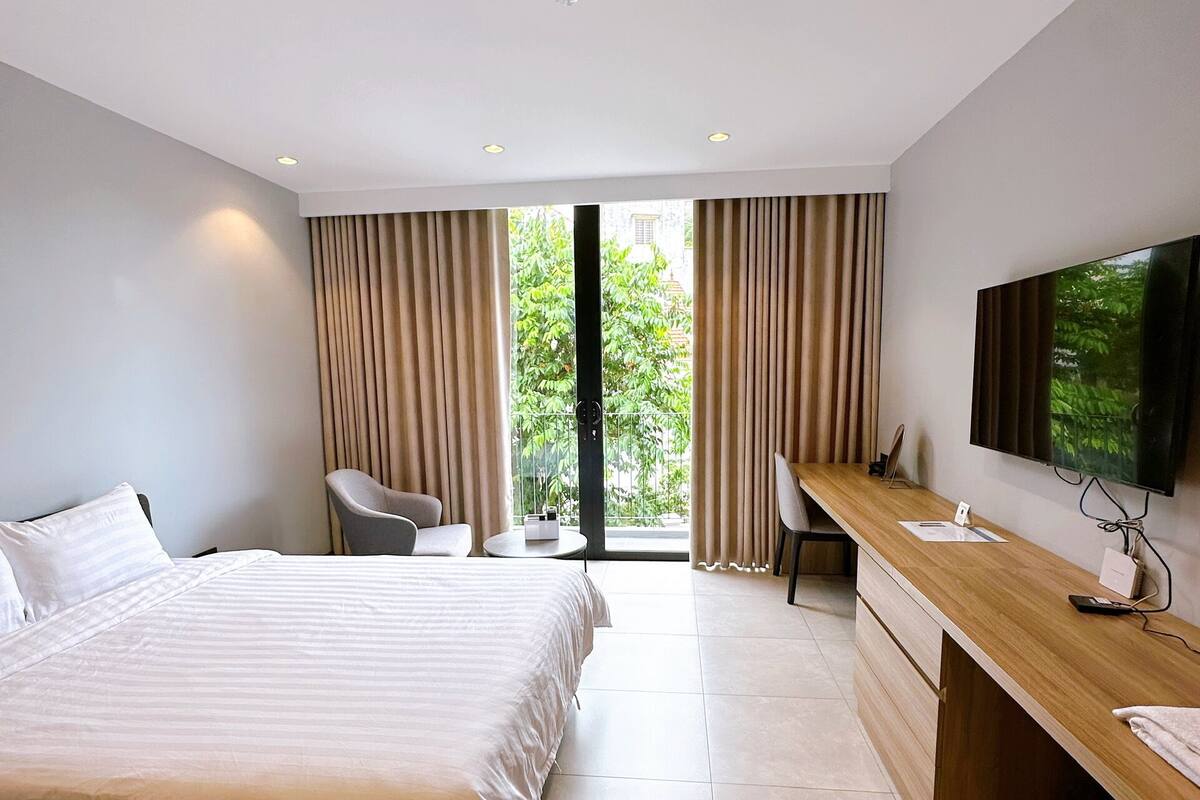 Ein modernes Hotelzimmer mit einem großen Bett, einem Schreibtisch, einem Flachbildfernseher, einem Sessel und Blick auf Grünflächen durch die Fenster.