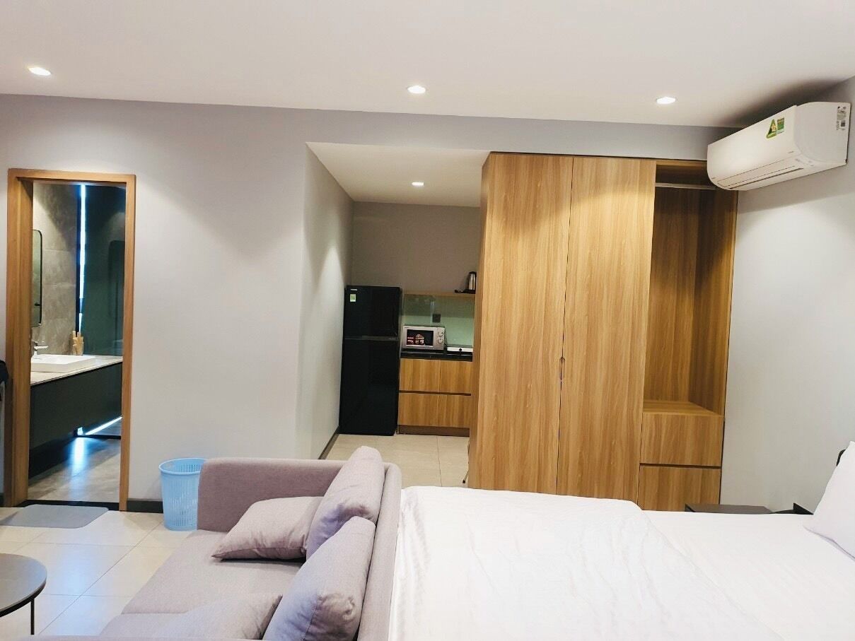 A modern hotel room with a bed, a seating area, a kitchenette, and a bathroom with a mirror and sink.