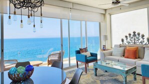 Una habitación con un ventanal grande con vista al mar, un sofá azul, un sofá blanco, una mesa de centro de cristal, sillas de mimbre y un candelabro.