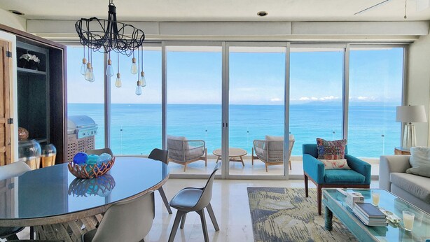Un balcón amplio con vistas al océano, una zona de comedor con mesa de vidrio y un espacio de descanso con sillas y sofá.