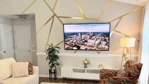 A modern living room with a flat-screen TV mounted on the wall, a white sofa, a colorful armchair, a small coffee table, and a potted plant. - Modern 3 BR 2 BA Home with Backyard – 8mi Downtown  (Charlotte)