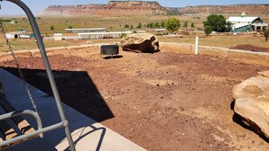 A view of a playground with a swing set and a sandbox, with a dirt ground and a building in the background. - The Rocky Mountain Cabin (Hurricane)