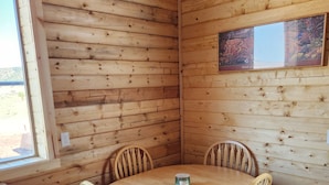 A wooden cabin interior with a round wooden table and four chairs, a window, and a painting on the wall. - The Rocky Mountain Cabin (Hurricane)