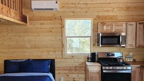 A log cabin interior with a bed, kitchenette, and a window with a view of trees. - The Rocky Mountain Cabin (Hurricane)