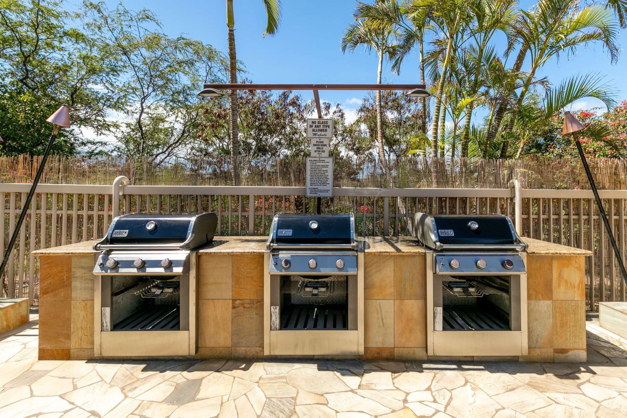 An outdoor barbecue area with three grills, a wooden fence, and a sign prohibiting glass containers.