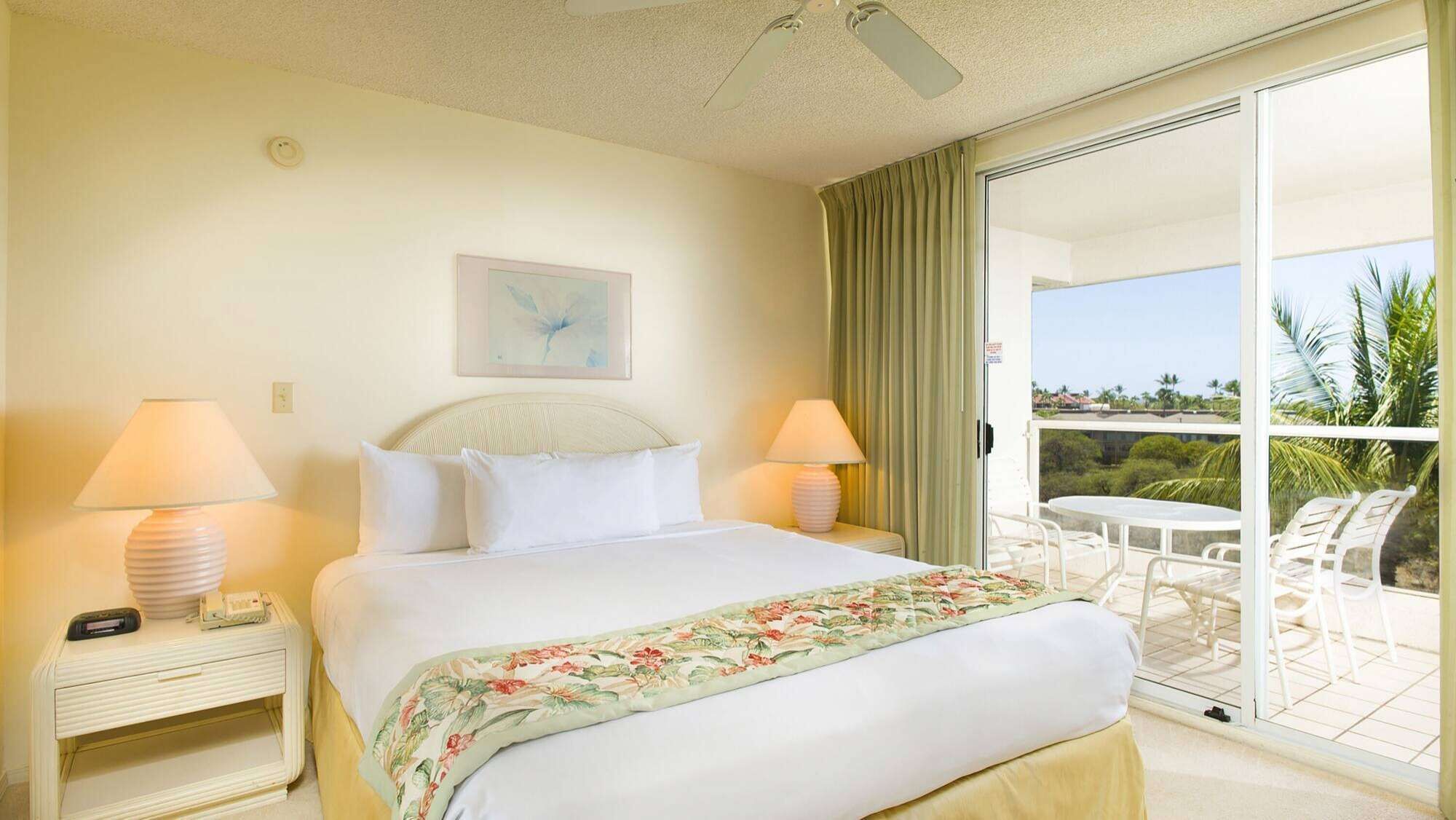 A hotel room with a bed, two bedside lamps, a nightstand, a window with curtains, and a balcony with a table and chairs.