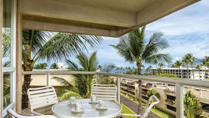 A balcony with a table and chairs, overlooking a parking area and palm trees. - Maui Banyan - 1BR Partial Ocean View - No Cleaning Fee/ Full Kitchen (Kihei)