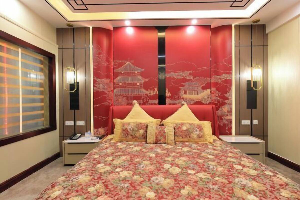 A bedroom with a bed, two nightstands, a window with blinds, and a red wall with a traditional building mural.