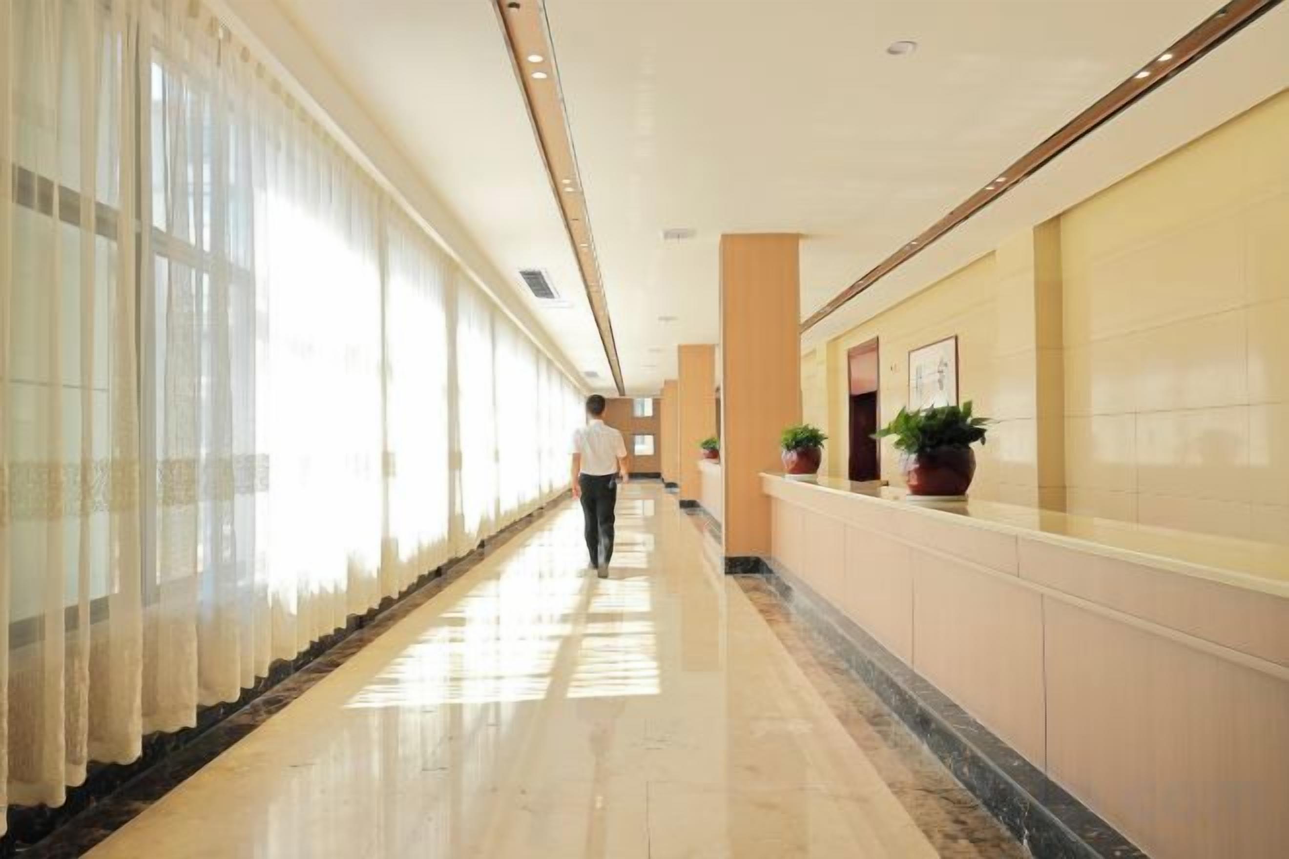 A long, well-lit hallway with a marble floor, large windows, and potted plants along the walls.