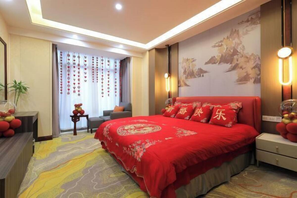 A hotel room with a large bed, a desk, a chair, a small table with decorative items, a potted plant, and a window with curtains.