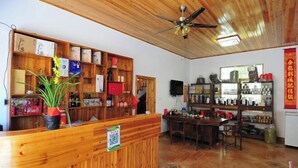 A wooden reception desk with a plant, shelves with various items, a ceiling fan, and a dining area with chairs and a table. - Yunshui Jingxiu Inn (Nanjing County)