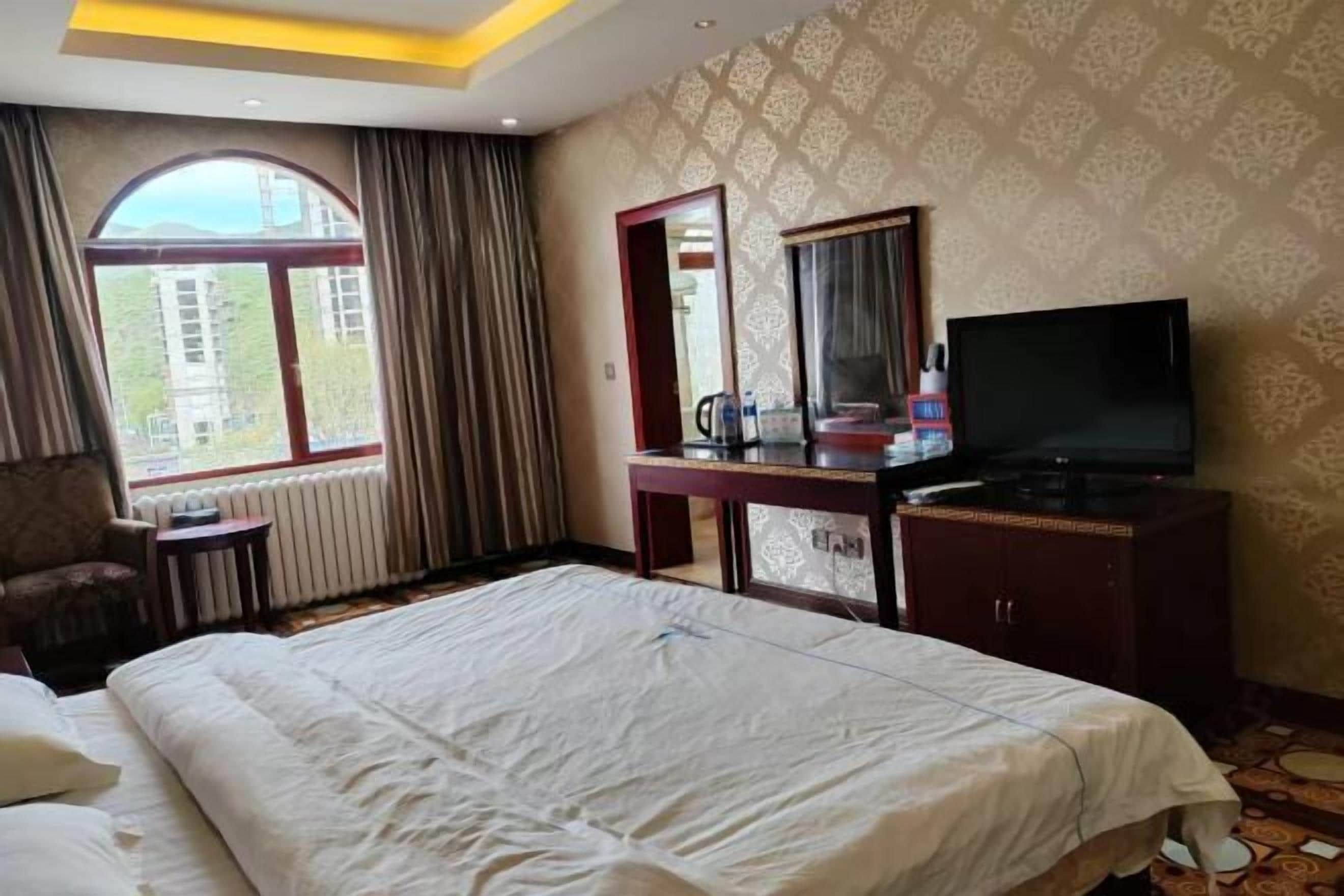A hotel room with a large bed, a desk with a TV, a chair, a window, and a mirror.