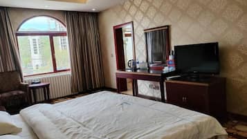 A hotel room with a large bed, a desk with a TV, a chair, a window, and a mirror.
