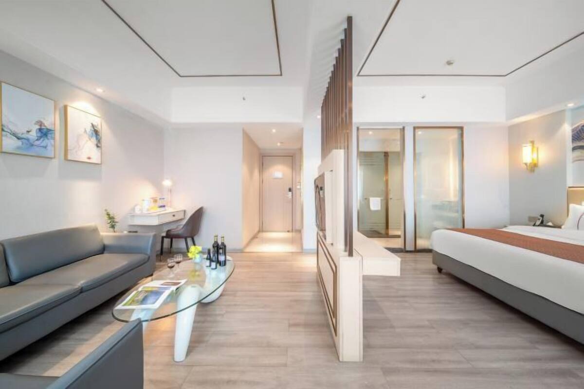 A modern hotel room with a sofa, a glass coffee table, a bed, and a desk with a chair.