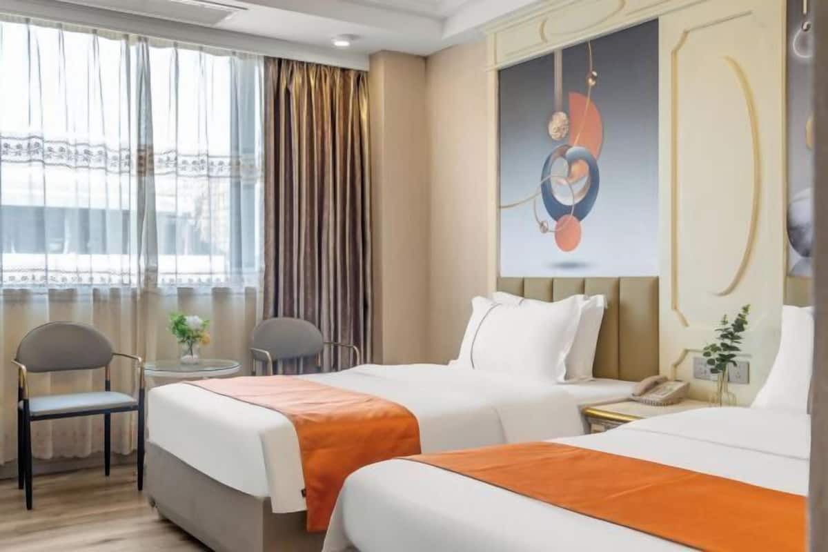 A hotel room with two beds, a small table, two chairs, a painting on the wall, and a window with curtains.