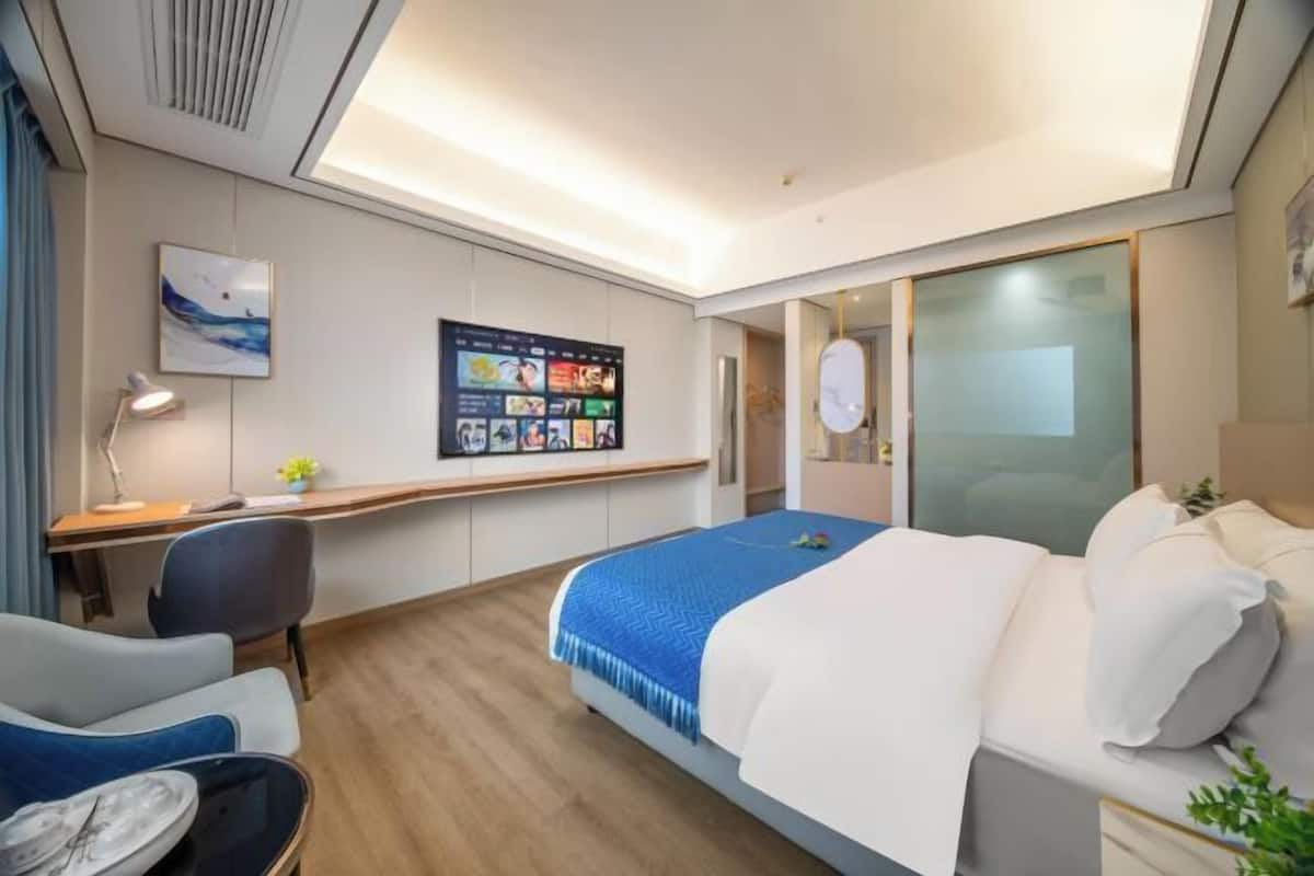 A modern hotel room with a bed, desk, chair, and a large digital screen displaying content.