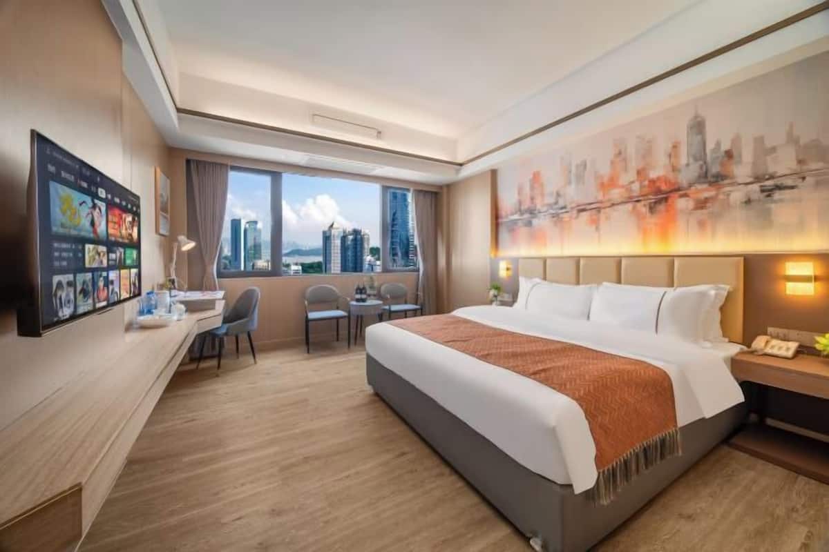 A hotel room with a large bed, a desk with a chair, a television mounted on the wall, and a cityscape view through the window.