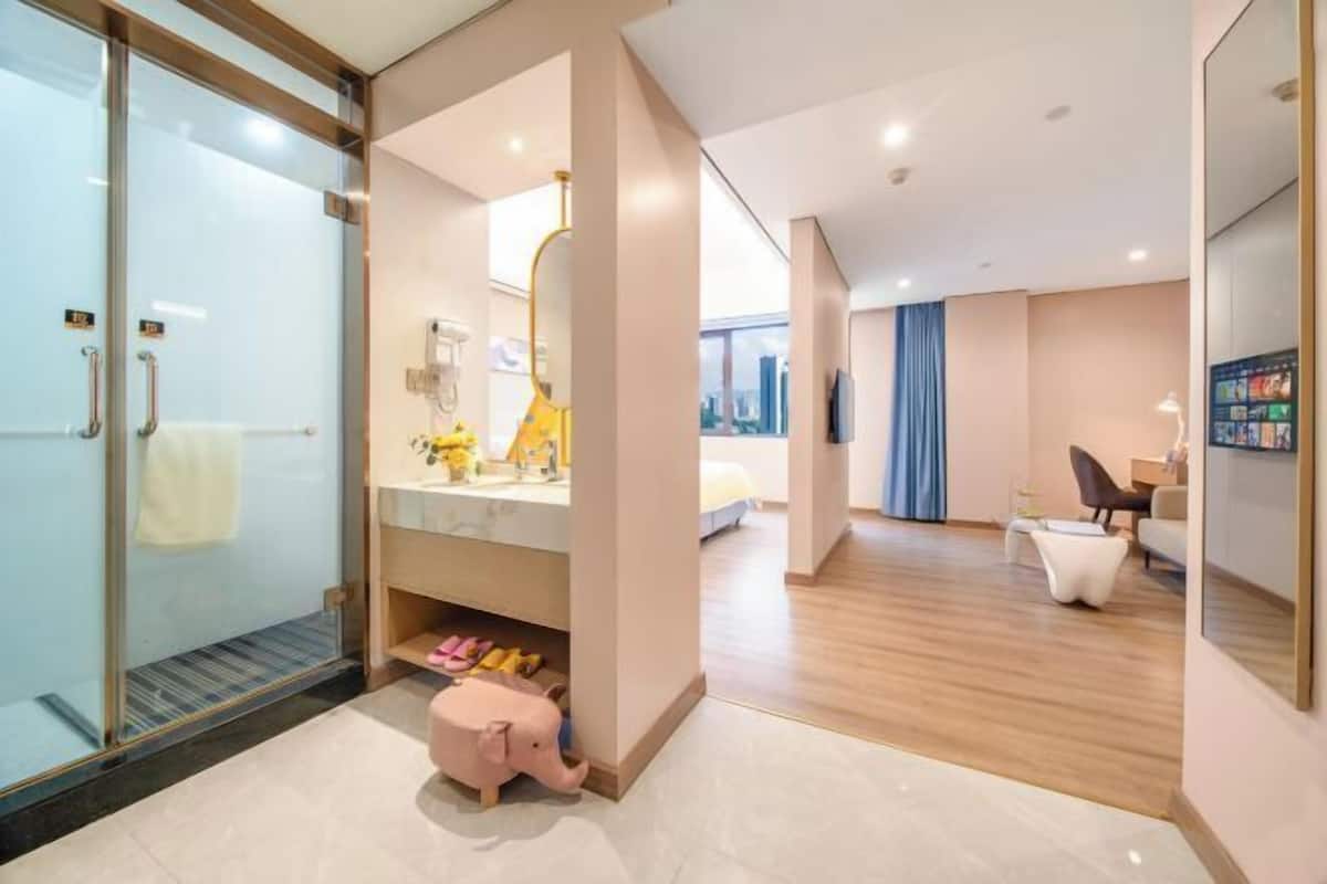 A modern hotel room with a glass shower enclosure, a vanity with a mirror, a bed with a city view, and a seating area with a table and chairs.
