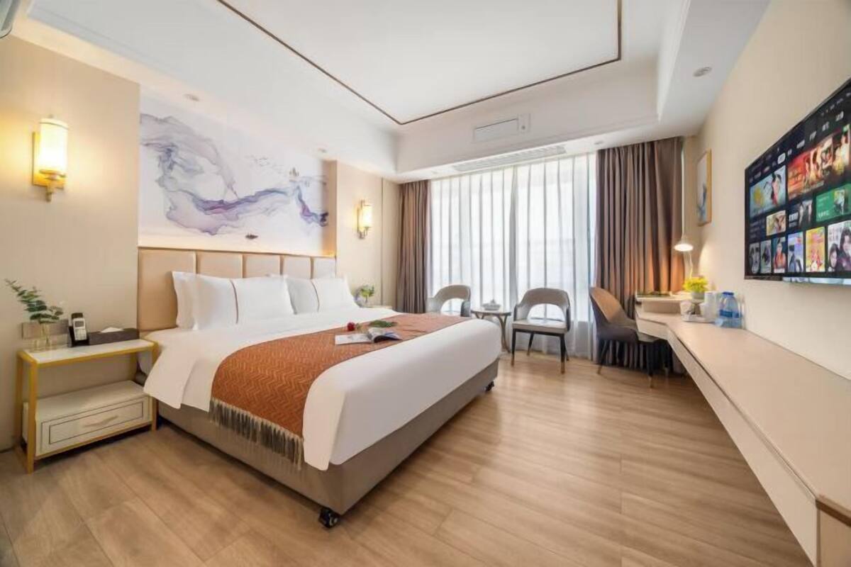 A modern hotel room with a large bed, bedside tables, a desk, and a large window with curtains.