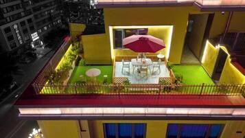 Un área de comedor al aire libre, bien iluminada, con una mesa y sillas, rodeada por un edificio amarillo con techo rojo.