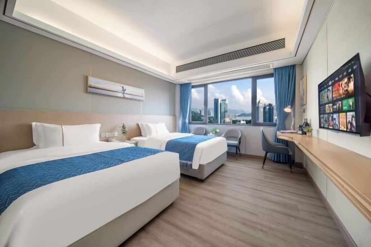 A hotel room with two beds, a desk, a chair, and a large window with a city view.