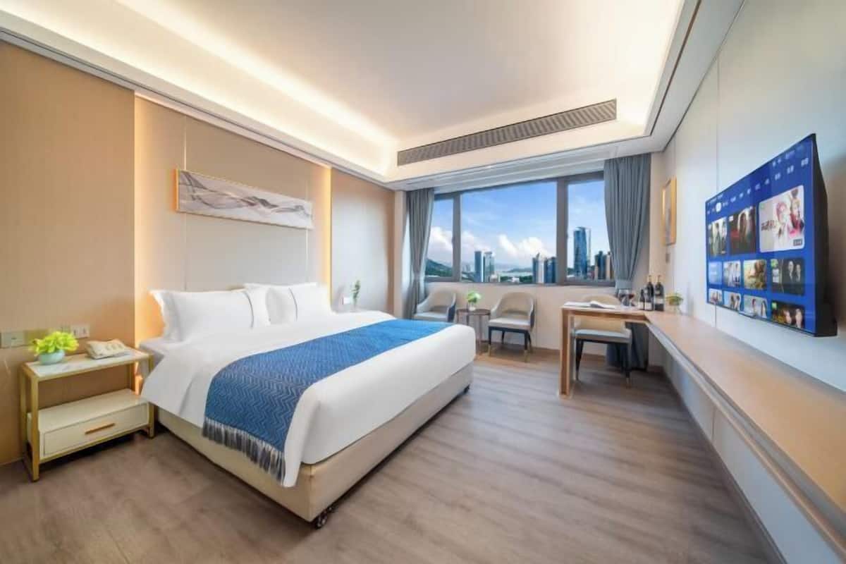 A hotel room with a bed, desk, chairs, a city view, and a wall with pictures.