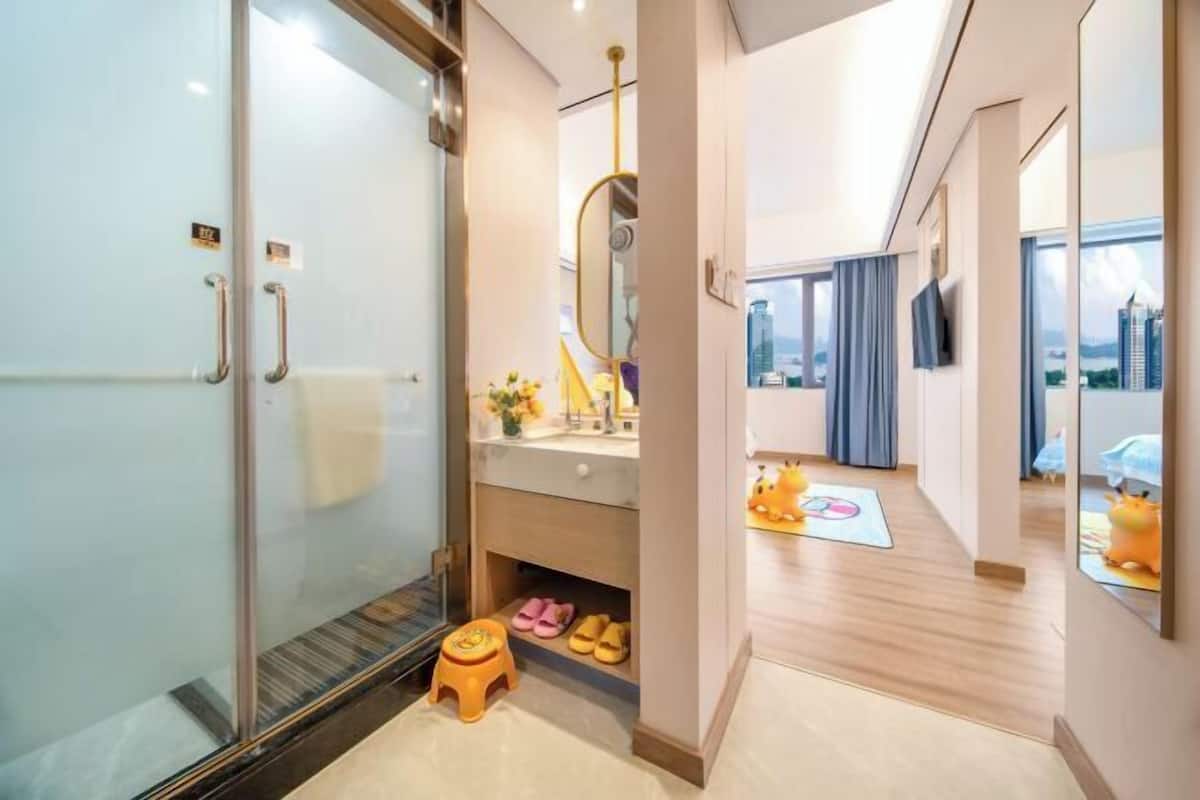 A modern hotel room with a glass shower enclosure, a child's play area, a large mirror, and a city view.