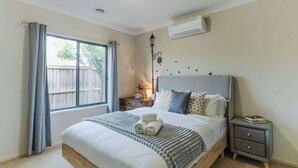 A neatly made bed with a gray headboard, a bedside table with a book, and a window with curtains. - Elegant 4BR Villa with 3 Living Rooms in PointCook (Point Cook)