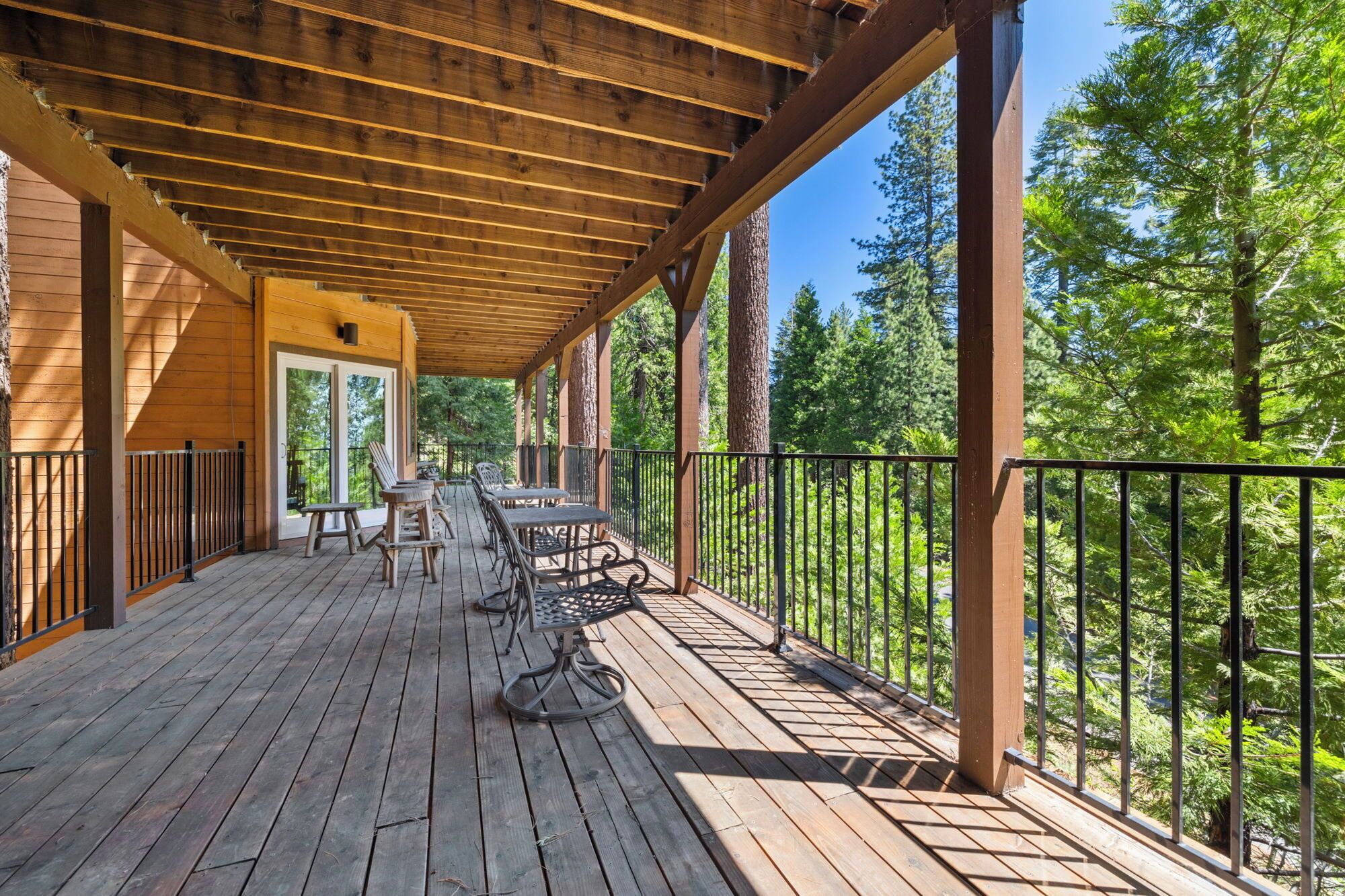 A wooden deck with outdoor seating, surrounded by a metal railing, overlooking a forested area.