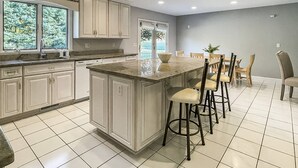 A kitchen with white cabinets, a large island with bar stools, and a view of trees through the window. - Akasa Ocean Front Oasis- Seasonal Pool, Grill (Stratford)