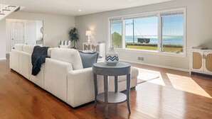 A modern living room with a white sofa, a round side table, and a view of the ocean through large windows. - Akasa Ocean Front Oasis- Seasonal Pool, Grill (Stratford)