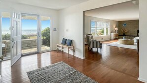 A spacious living room with a wooden floor, a bench with cushions, and a view through glass doors to the outdoors. - Akasa Ocean Front Oasis- Seasonal Pool, Grill (Stratford)