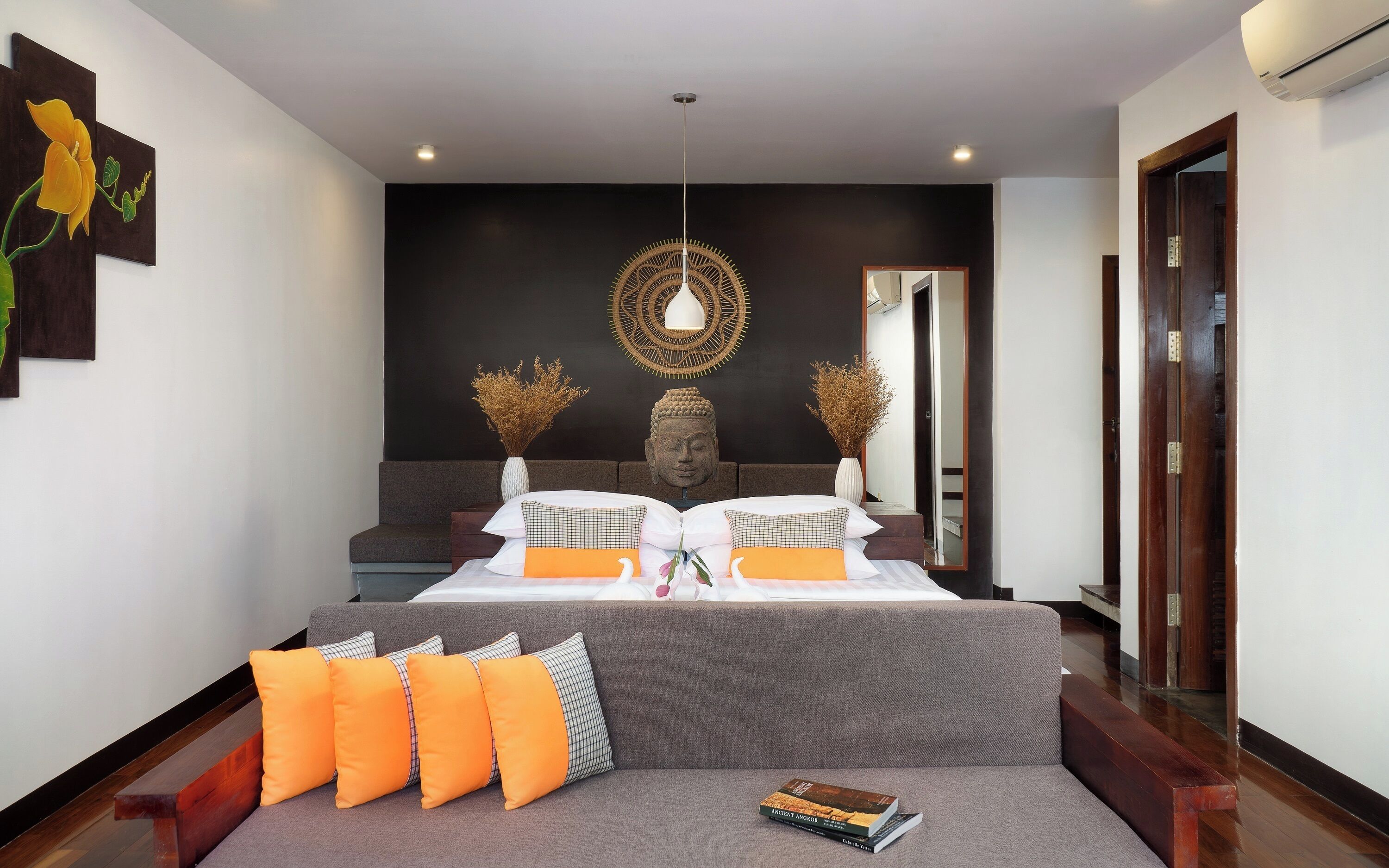 A modern bedroom with a grey sofa, a large bed with white and orange bedding, a Buddha head sculpture, and a wall-mounted light fixture.