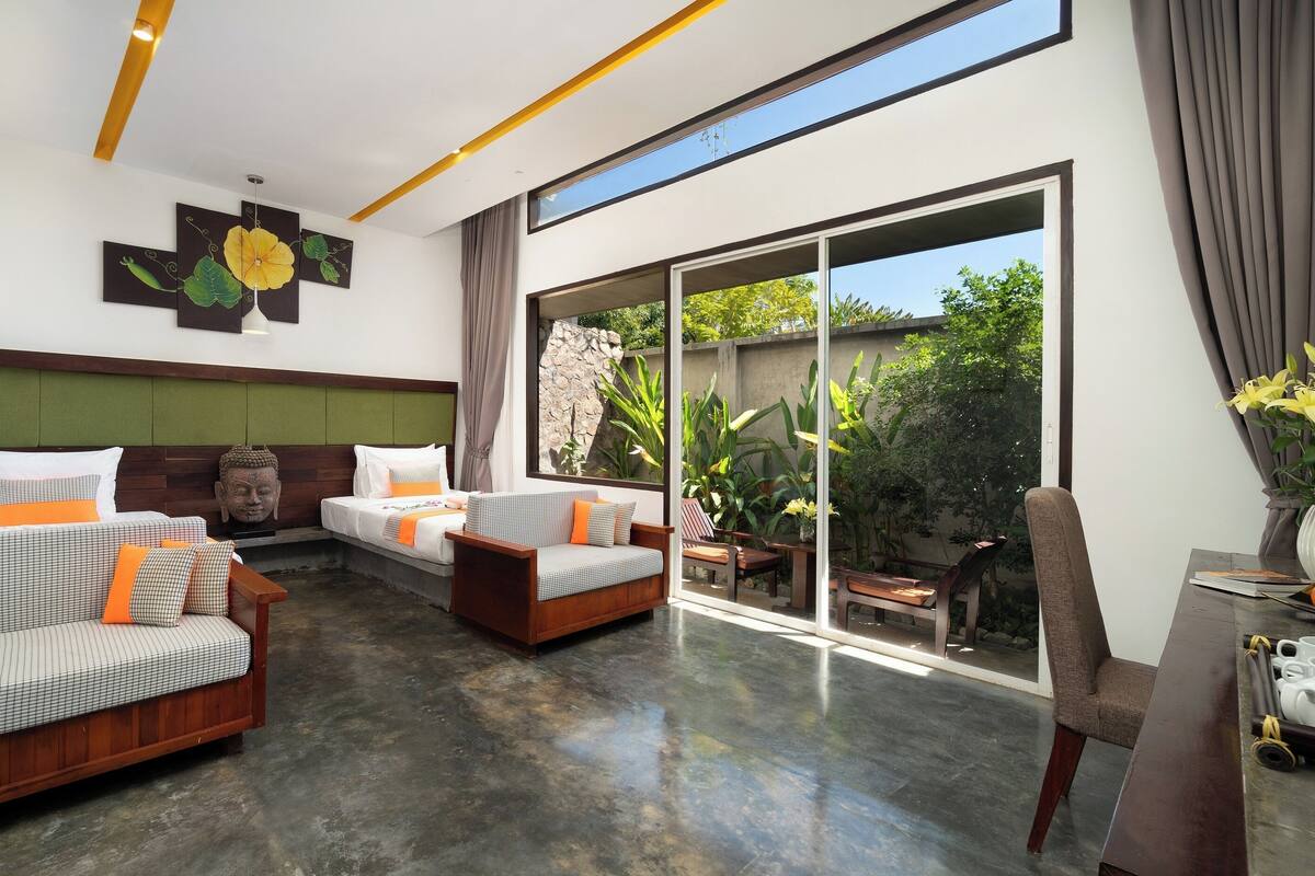 Camera d'albergo moderna con un'ampia porta scorrevole in vetro che si apre su un giardino, una divano letto, una testiera in legno e una statua di Buddha.