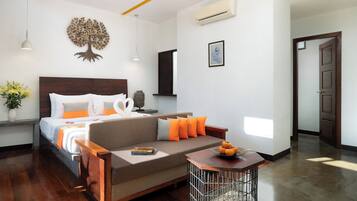 A modern hotel room with a wooden headboard, a bed with white and orange bedding, a grey sofa with orange cushions, a small wooden table with a bowl of fruit, and a Buddha statue.