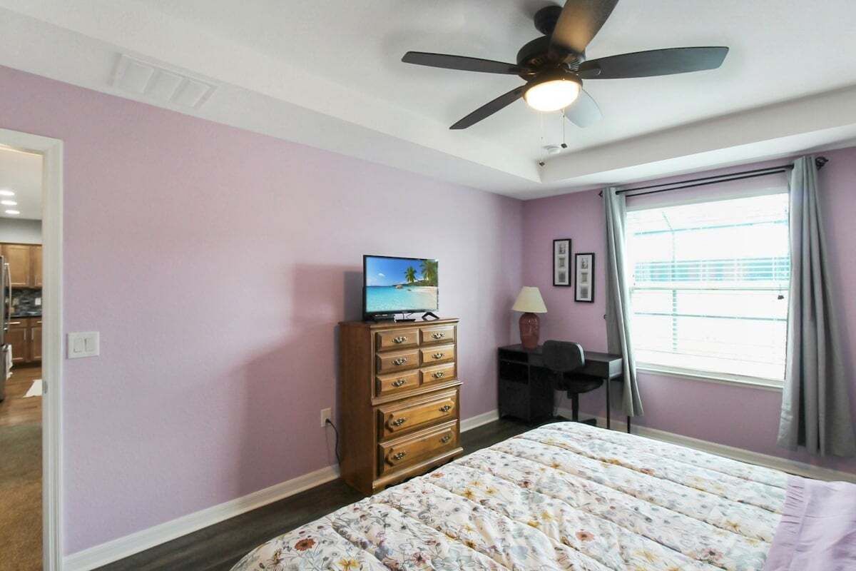 A bedroom with a bed, a dresser, a desk, a ceiling fan, and a window with curtains.