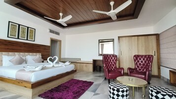 Ein Hotelzimmer mit einem Bett, zwei roten Sesseln, einem kleinen Tisch, einem violetten Teppich und Deckenventilatoren.