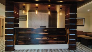 A reception desk with a modern design, featuring a wooden panel with a unique pattern and a white base. There are three pendant lights above the desk, and the area is surrounded by dark wooden panels. - Vbrance - Luxury Boutique Hotel (Puri)