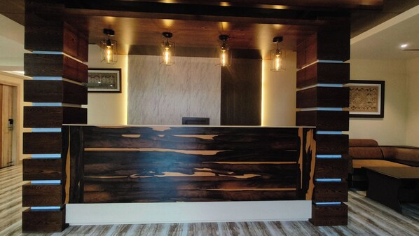 Een receptiebalie met een modern ontwerp, met een houten paneel met een uniek patroon en een wit basisstuk. Daarboven hangen drie hanglampen, en de ruimte wordt omringd door donkere houten panelen.