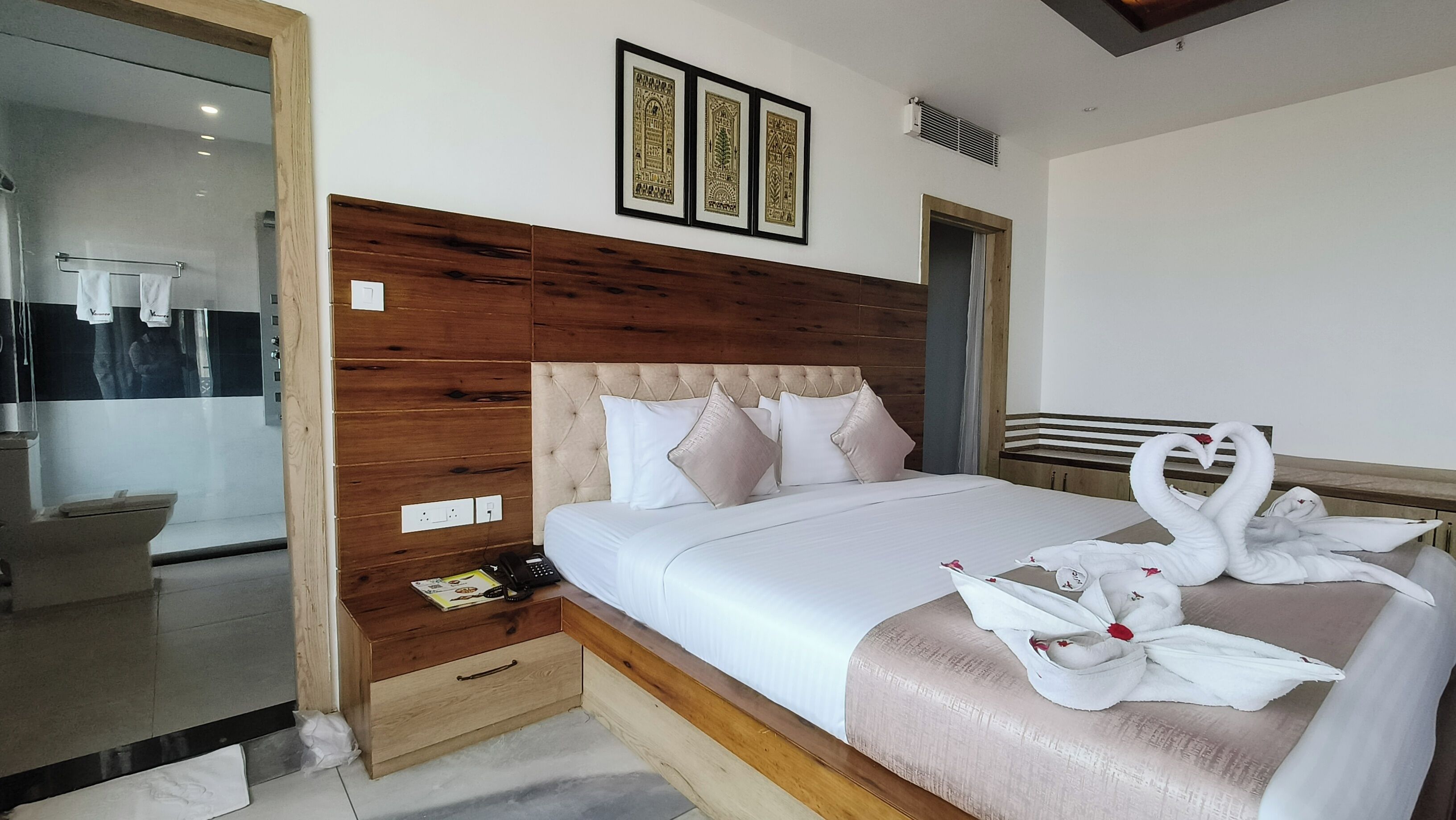Une chambre d’hôtel avec une tête de lit en bois, un lit avec une literie blanche, une table de chevet avec un téléphone et un magazine, et un miroir avec un porte-serviettes.