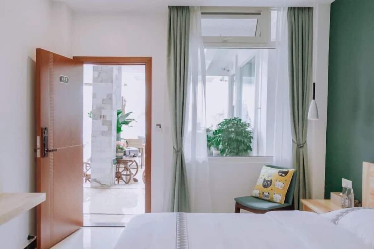 A hotel room with a bed, a chair with a cat-themed pillow, a window with curtains, and a door leading to a balcony.