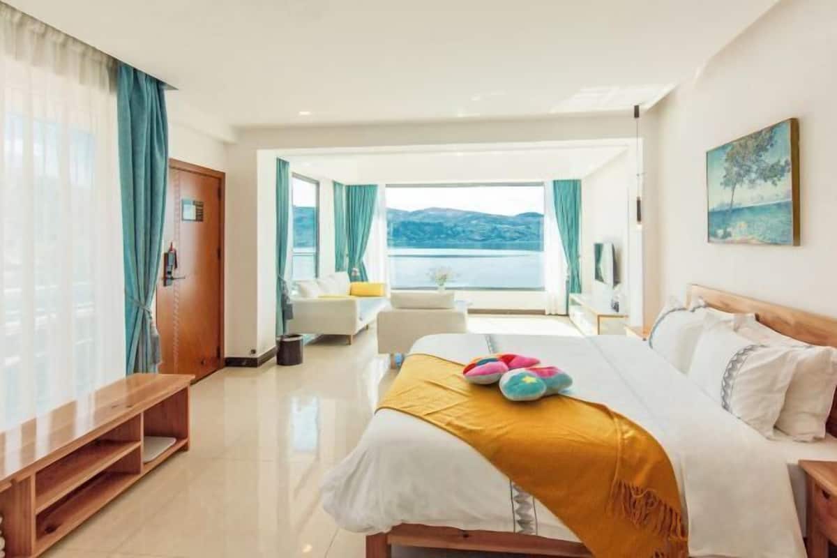A hotel room with a bed, a wooden bench, a view of the sea, and a painting on the wall.