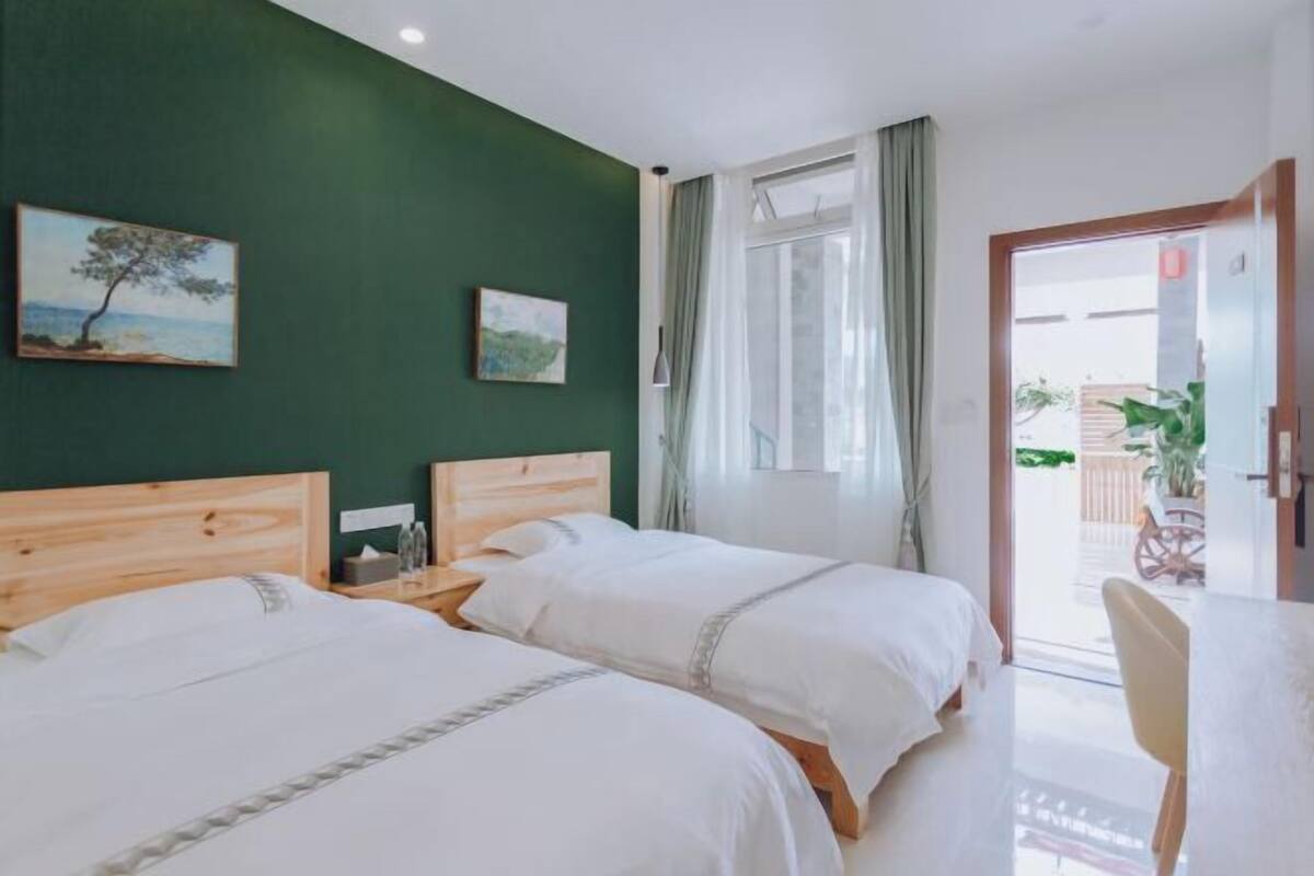 A room with two beds, a wooden headboard, a small table with a chair, and a view of the outside through a door.