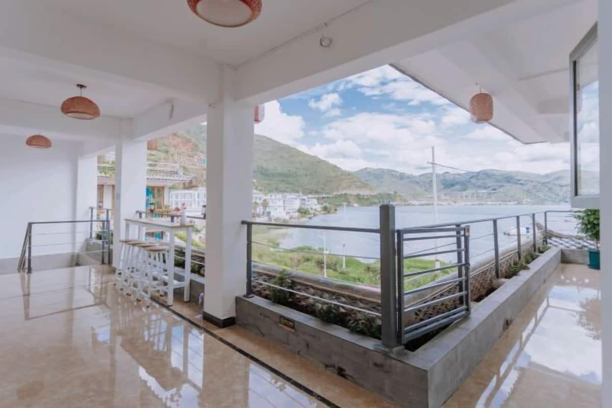 A spacious indoor area with a view of a lake and mountains. It features a polished floor, a railing overlooking the water, and hanging pendant lights.