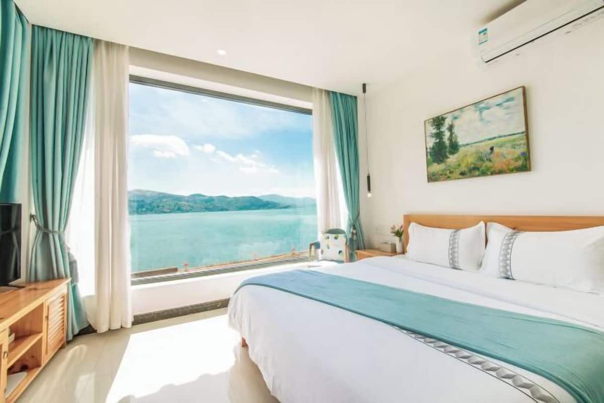 A hotel room with a large window offering a view of a lake and mountains, a bed with white bedding and a teal throw, and a wooden nightstand with a television.