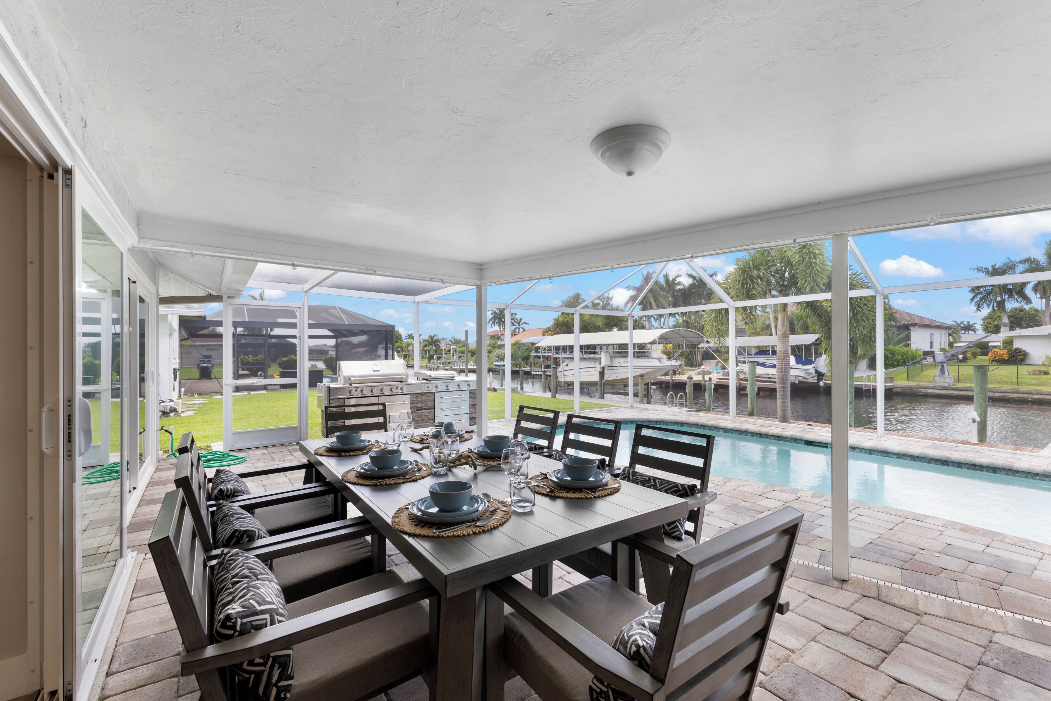 A covered patio with a dining table set for six, overlooking a swimming pool and a marina.