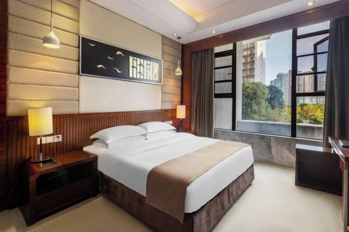 Habitación de hotel con una cama grande, una mesita de noche, una obra de arte colgada en la pared, una ventana con vista a la ciudad y un cabecero de madera.