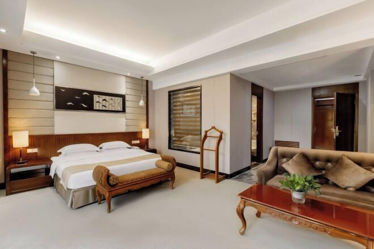 Una habitación de hotel con cama, mesitas de noche, sofá, mesa de centro y un ventanal grande.
