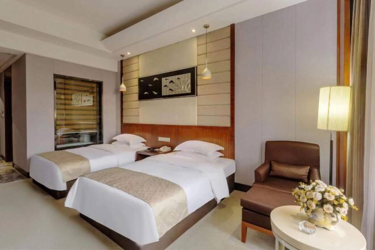 Habitación de hotel con dos camas, un sillón marrón, una mesita de noche con lámpara y una obra de arte colgada en la pared.